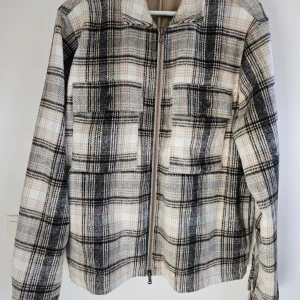 Rutig skjortjacka från Riley i ullmix - Snygg rutig skjortjacka från Riley i beige, svart och grå toner. Jackan har dragkedja framtill, två bröstfickor med knappar och klassisk krage. Materialet är en mjuk ullmix som passar perfekt för lager-på-lager-stil.