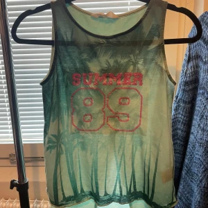 Grön linnetopp med palmmotiv H&M - Cool ärmlös linnetopp från H&M i gröna nyanser med palmmotiv och texten SUMMER 89 i glittriga rosa bokstäver. Toppen har omlottöppning baktill för en extra snygg detalj. Perfekt för varma dagar och somrig vibe. Barntröja men passar även mindre vuxna. 