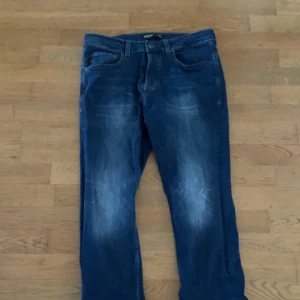 Blå jeans från Burton London  - Säljer ett par klassiska blå jeans från Burton London med snyggt slitna detaljer. Jeansen har fem fickor, knappgylf och är tillverkade i robust denim. Perfekt för dig som gillar en tidlös och avslappnad stil.
