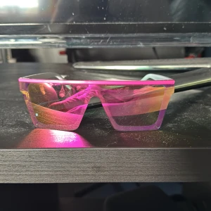 Rosa och vita futuristiska solglasögon - Säljer ett par riktigt coola solglasögon med rektangulär form och breda linser. Glasögonen har en stark rosa front och vita skalmar, vilket ger en futuristisk vibe. Perfekt för dig som vill sticka ut och addera färg till din stil. Materialet är plast.