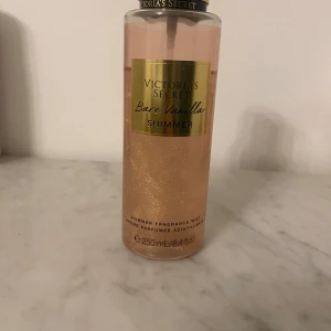 Victoria's Secret Bare Vanilla Shimmer - Säljer en Victoria's Secret Bare Vanilla Shimmer body mist med doft av vispad vanilj och mjuk cashmere. Flaskan är genomskinlig med guldiga detaljer och innehåller 250 ml skimrande mist som ger huden en glittrande finish.