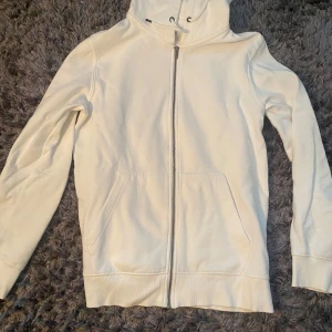 Zip hoodie från jack&jones  - Säljer en stilren vit hoodie med hel dragkedja framtill och praktisk huva. Tröjan har långa ärmar och två fickor på magen. Perfekt för chill dagar eller när du vill ha en enkel look. Passar dig som gillar basic och cleana plagg.