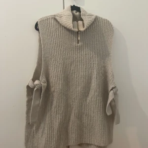 Beige stickad väst med halv dragkedja - Säljer en beige stickad väst med hög krage och halv dragkedja framtill. Västen har breda sidoslitsar med knytband i sidorna och en liten metallknapp med ananasdetalj. Perfekt för lager-på-lager och chill stil.