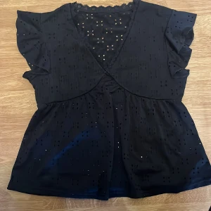 Svart blus med spets från SHEIN - Svart blus från SHEIN med v-ringning och spetsdetaljer längs halsringningen. Blusen har korta volangärmar och är dekorerad med små hålbroderier som ger en luftig känsla. Tillverkad i polyester med lite stretch för extra komfort.