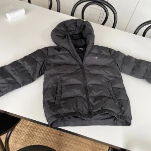 Svart soft dunjacka från gant - Svart pufferjacka från GANT med huva och dragkedja framtill. Jackan har ett stilrent, quiltat mönster och två sidofickor. Perfekt för kalla dagar med sin fluffiga och bekväma passform. Diskret GANT-logga på bröstet och elastiska muddar vid ärmslut. Använd max 10 gånger 