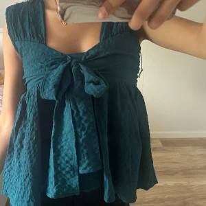 Turkos blus med stor rosett - Supersöt turkos blus med breda axelband och en stor knytrosett framtill. Storlek XS/S