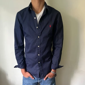 Mörkblå skjorta från Ralph Lauren - Snygg mörkblå skjorta från Ralph Lauren med klassisk krage och knappar hela vägen. Broderad röd logga på bröstet ger en stilren touch. Skjortan är långärmad och gjord i mjuk bomull som känns skön mot huden. Perfekt för dig som gillar enkel och tidlös stil. Skriv frågor ifall ni har några funderingar.