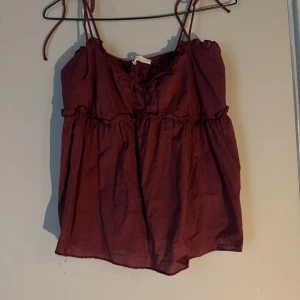 Vinrött linne/topp med volangdetaljer - Säljer en vinröd linne/topp med tunna axelband som man knyter och volangdetaljer både upptill och i midjan. Toppen har en luftig och flowy passform 🥰