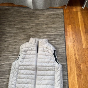Ljusgrå quiltad dunväst - Snygg ljusgrå dunväst med quiltad design och hög krage. Västen har en hel dragkedja framtill och är perfekt att slänga över en hoodie eller tröja när det är lite kyligt ute. Den har en clean look utan synliga loggor eller mönster. Storlek S.