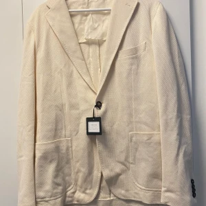 Tagliatore Beige Kavaj - Snygg beige kavaj med strukturerat tyg och klassisk krage. Kavajen har två stora framfickor, en bröstficka och fyra svarta knappar vid ärmslut. Passformen är normal och tyget känns som bomull eller linne. Perfekt för dig som gillar stilrena och ljusa plagg.