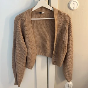 Beige croppad kofta från H&M Divided - Säljer en beige croppad kofta från H&M Divided i storlek XS. Koftan är stickad med grov ribbad struktur, har långa ärmar och tre stora knappar framtill. Perfekt för lager-på-lager och chill stil. Passar dig som gillar loose fit och neutrala färger. Tröjan har en liten tråd som sticker ut i armen som du kan se på bilden men det ska inte vara något problem att bara väva in den igen!❤️