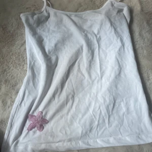 Vit linnetopp med rosa blomdetalj - Säljer en vit linnetopp med smala axelband och en söt rosa blomma i pärlor nere vid kanten. Enkel och fräsch design som passar perfekt till sommaren. Tillverkad i mjuk bomull för en skön känsla. Gjort själv