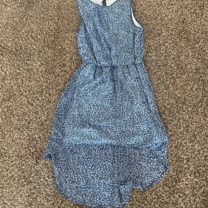 Blommig blå klänning från H&M - Snygg ärmlös klänning från H&M med blått blommigt mönster och rund hals. Klänningen har en asymmetrisk nederdel som är längre bak och ett mjukt, lätt material som känns skönt mot huden. 