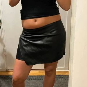 Svart skinnkjol - Snygg svart kort skinnkjol med omlott-look. Kjolen har en stilren design och sitter tajt i midjan. Perfekt för dig som vill ha en edgy vibe och gillar minimalistiska plagg med attityd. Inbyggda skjorts i kjolen