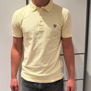 Gul piké från Lyle & Scott, storlek M - Snygg ljusgul pikétröja från Lyle & Scott med klassisk krage och tre knappar framtill. Ikonisk logga på bröstet. Tillverkad i mjuk bomull som känns skön mot huden. Perfekt för dig som gillar stilren och enkel design.