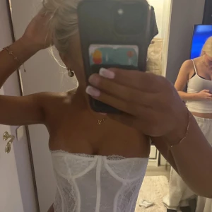 Vit spetskorsett med bandeau-stil - Säljer en superfin vit korsett i bandeau-modell med spetsdetaljer och markerade sömmar framtill. Toppen är axelbandslös och har en snygg, figurnära passform som ger en riktigt trendig look. Perfekt att styla med jeans eller kjol för en chic vibe.