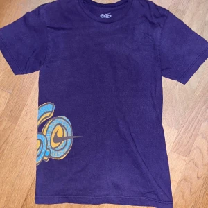 Lila Nike t-shirt med grafiskt tryck - Cool lila t-shirt från Nike med korta ärmar och ett fett grafiskt tryck i blått och gult på både fram- och baksidan. Trycket har en streetstyle-vibe och t-shirten är gjord i mjuk bomull. Perfekt för dig som gillar sportiga och unika plagg.