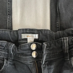 Mörkgrå jeans från Gina Tricot - Snygga mörkgrå jeans från Gina Tricot med raka ben och klassisk femficksdesign. Jeansen har dubbla silverknappar i midjan och dragkedja, samt bakfickor med knappdetalj. Perfekta för en chill och trendig look.
