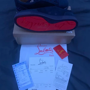 Svarta sneakers från Christian Louboutin - Snygga svarta sneakers från Christian Louboutin med klassisk röd sula och diskret logga på sidan. Skorna har snörning och är tillverkade i mocka med läderdetaljer. Perfekta för dig som vill ha en lyxig och stilren look med en ikonisk touch. Box hörn är av har fixat den dock och 1100kr prutat å klart 