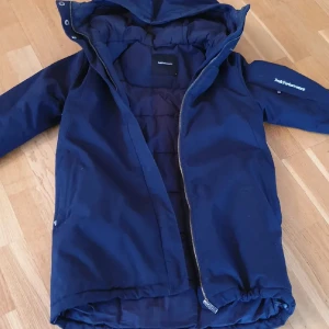 Blå dunjacka från Peak Performance - Peak Performance dunjacka i mörkblått med huva och dragkedja. Jackan har en sportig look, är vadderad och har logga på ärmen. Perfekt för kalla dagar och har en klassisk passform med långa ärmar.