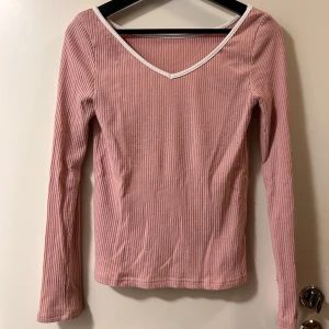 Rosa ribbad långärmad topp - Säljer en ljusrosa ribbad topp med lång ärm och v-ringad hals som har vit kant. Toppen är slim fit och har en mjuk, stretchig känsla. Perfekt för dig som gillar enkel men snygg stil med lite extra detalj vid halsen.