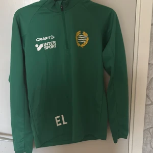 Grön Hammarby Craft träningströja - Säljer en grön långärmad träningströja från Craft med Hammarby IF-emblem på bröstet och halv dragkedja. Tröjan har tryck från Intersport och initialerna EL framtill. Perfekt för fotbollsträning eller sportiga dagar.