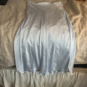 Ljusblå långkjol från The Black Label - Säljer en ljusblå långkjol från The Black Label Collection, modell STEPHIE. Kjolen har en mjuk, glansig yta och faller snyggt längs benen. Perfekt för dig som gillar stilrena och eleganta plagg med flow. Materialet känns svalt och lyxigt.