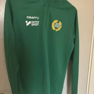 Grön Hammarby Craft half zip tröja - Grön långärmad träningströja från Craft med Hammarby IF-logga och Intersport-tryck på bröstet. Tröjan har en praktisk half zip-dragkedja vid halsen och är gjord i ett mjukt, sportigt polyestermaterial. Perfekt för fotbollsträning eller chill.
