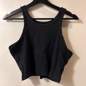 Svart ribbad croppad linnetopp - Säljer en svart croppad linnetopp med ribbad struktur. Toppen är ärmlös och har en hög halsringning, perfekt för att styla med höga jeans eller kjol. Enkel och stilren design som funkar till många olika looks.