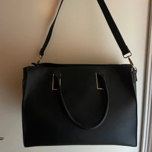 Svart axelväska i skinnimitation - Stilren svart axelväska med både handtag och axelrem. Väskan har guldfärgade metalldetaljer och dragkedja upptill. Rymmer mycket och har en klassisk, boxig form. Perfekt för dig som vill ha en snygg och praktisk väska till vardagen. Båda sidor går även att knäppa. Pris går att diskuteras.