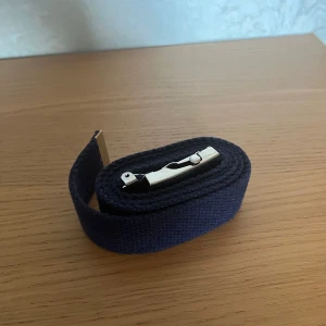 Mörkblå canvasbälte med metallspänne - Snyggt mörkblått bälte i kraftig canvas med ett stilrent, silvrigt metallspänne. Bältet har en enkel och clean design utan mönster. Metallspännet är robust och lätt att justera för bästa passform. 100 cm lång.