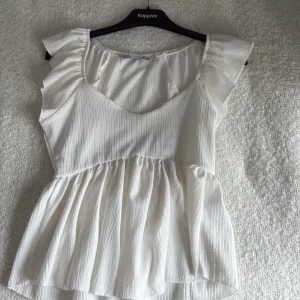 Vit volangblus från 157, S - Supersöt vit blus från 157 med volangärmar och v-ringad hals. Toppen har en lätt rynkad, flowy nederdel och är gjord i ett ribbat tyg som ger en snygg struktur. Perfekt för dig som gillar romantiska och luftiga plagg.