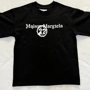 Maison Margiela svart t-shirt med tryck - Svart t-shirt från Maison Margiela med vit logotyp och grafiskt tryck framtill. Baksidan har ett coolt kalendertryck i vitt. Klassisk rund hals och korta ärmar. Materialet är mjuk bomull, perfekt för dig som gillar statement-plagg med unik design.