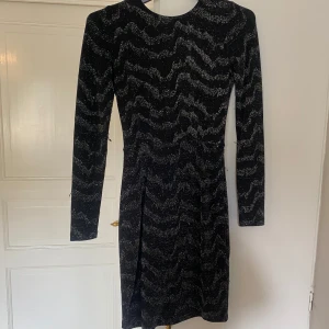 Glittrig svart klänning Gina Tricot XS - Svart kortklänning från Gina Tricot i storlek XS med glittrigt mönster och lång ärm. Klänningen har öppen rygg med rosett och en tight passform. Några trådar som sticker ut men inga andra defekter. Hör av er vid frågor!💕
