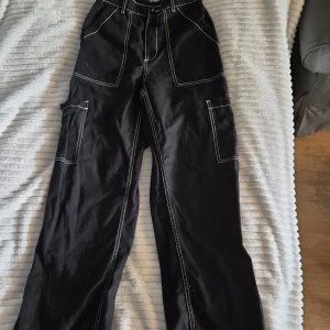Svarta cargopants från H&M Divided - Svarta cargopants från H&M Divided med kontrasterande vita sömmar. Byxorna har stora fickor på sidorna och bak, samt bred midja med hällor. Tillverkade i bomull för en skön och avslappnad känsla. Perfekta för en streetwear-look.