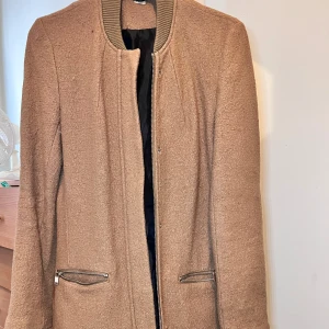 Beige ullkappa med dragkedjor - Stilren beige kappa i ullmix med raka linjer och ribbad krage. Jackan har två fickor med silvriga dragkedjor och svart foder på insidan. Perfekt för dig som gillar en clean look och vill hålla dig varm under höst och vinter.