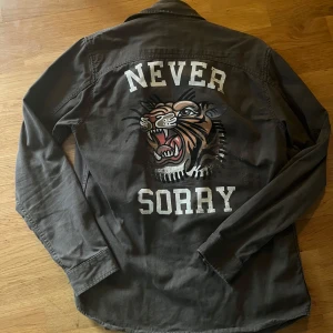Grön skjorta Jack & Jones - Snygg grön skjorta/overshirt med coolt tigertryck och texten 'NEVER SORRY' på ryggen. Använd fåtal gånger, mycket bra skick!
