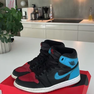Nike Air Jordan 1 High OG unc to chi - Nike Air Jordan 1 High OG i svart, rött och blått skinn. Klassisk high-top siluett med svart snörning, röd tåbox och yttersula, blå detaljer på häl och swoosh. 