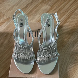Glittriga silvriga skor från Truffle - Snygga skor i metallic silver från Truffle med glittriga strassdetaljer över foten. Skorna har öppen tå, smal rem runt hälen och låg klack (5-6 cm). Perfekta för dig som vill sticka ut med extra bling och glam på fötterna.
