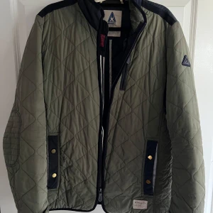 Quiltad grön jacka från GAASTRA - Snygg quiltad jacka från Gaastra i olivgrönt med svarta detaljer på axlar och insida. Jackan har guldiga knappar, dragkedja och två fickor framtill. Perfekt för dig som gillar stilren och sportig look. Fodrad insida för extra komfort. Perfekt för kommande höst. Storlek XL men funkar även för L.