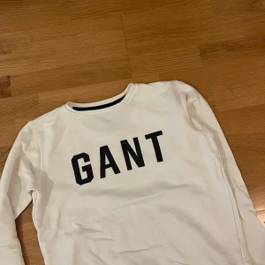 Vit sweatshirt från GANT med tryck - Vit sweatshirt från GANT med svart logotyptryck över bröstet. Klassisk rund halsringning och långa ärmar. Mjuk insida och ribbade muddar vid ärmslut och nederkant. Perfekt för en clean och stilren look.