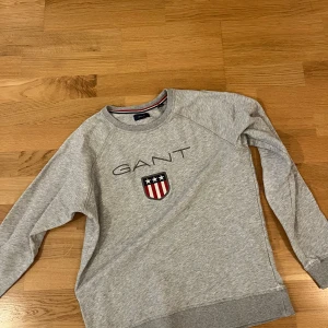 Grå sweatshirt från GANT med logo - Snygg grå sweatshirt från GANT med broderad logga och sköld på bröstet. Tröjan har rund hals, långa ärmar och ribbade muddar. Perfekt för dig som gillar klassisk och sportig stil. Passar dig som är ca 170 cm.