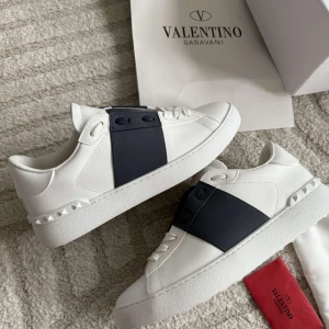 Valentino Garavani Open sneakers blå/vit - Snygga Valentino Garavani Open sneakers i vitt skinn med bred blå läderdetalj över vristen. Klassisk rund tå, vita snören och ikoniska nitar på hälen. Perfekt för dig som vill ha en clean men lyxig look.