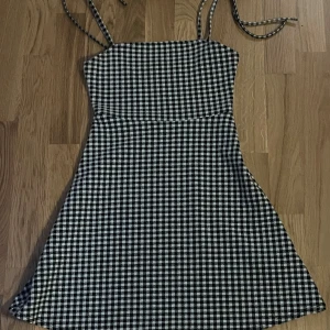 Svartvit rutig kortklänning - Säljer en svartvit rutig kortklänning med smala axelband och lätt utställd kjol. Klänningen har en klassisk fyrkantig urringning och är perfekt för dig som gillar en enkel men trendig stil. Materialet känns lätt och passar bra till varma dagar.