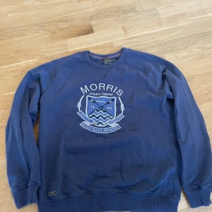 Blå sweatshirt från Morris, storlek L - Snygg blå sweatshirt från Morris med stort tryck framtill och klassisk collegekänsla. Tröjan har rund halsringning, ribbade muddar och är tillverkad i mjuk bomull. Perfekt för en avslappnad och stilren look.