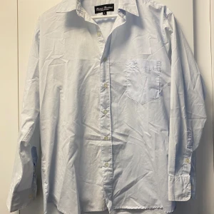 Ljusblå skjorta  - Snygg ljusblå skjorta från Roberto Bertolucci i storlek L. Klassisk modell med krage, bröstficka med broderad logga och knappar framtill. Tillverkad i mjuk bomull och har långa ärmar med manschett. Perfekt för dig som gillar stilrena och enkla plagg.