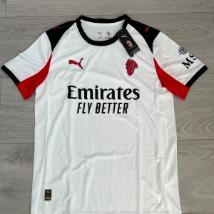 AC Milan vit fotbollströja - AC Milan fotbollströja från Puma i vit med svarta och röda detaljer. Kortärmad modell med svart krage och tryckta loggor på bröstet. 