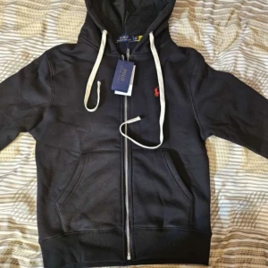 Ralph Lauren Zip Hoodie - Säljer en stilren svart hoodie från Ralph Lauren med dragkedja och vit snörning. Den har en röd broderad logotyp på bröstet och är perfekt för en avslappnad look. Helt ny med pris lapp och helt oanvänd! Skicka 10/10. Köp direkt eller ge prisförslag 💛 