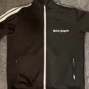 Svart träningsjacka från Palm Angels - Snygg svart träningsjacka från Palm Angels med vita ränder på ärmarna och dragkedja framtill. Jackan har en hög krage och två fickor med dragkedjor. Perfekt för en sportig look.