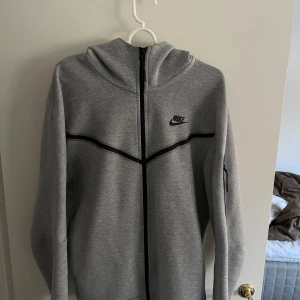 Nike Tech grå - Säljer en stilren grå hoodie från Nike med dragkedja och svart logotyp på bröstet. Tröjan har en huva och långa ärmar, perfekt för en avslappnad look. Den har också svarta detaljer som ger en sportig känsla. Skick 9/10, använd ett fåtal gånger. Storlek M